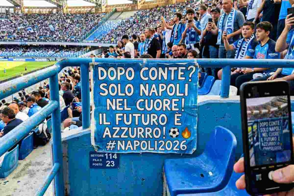 Foto realistica scattata da smartphone allo stadio Diego Armando Maradona di Napoli durante una partita: un tifoso in primo piano regge un cartello azzurro con la scritta bianca "DOPO CONTE? SOLO NAPOLI NEL CUORE! IL FUTURO È AZZURRO! #NAPOLI2026". Lo stadio è gremito di tifosi e la partita è in corso sul campo. Un altro tifoso scatta una foto al cartello con il proprio telefono.