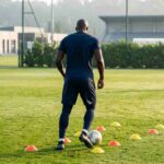 Un calciatore che sembra Romelu Lukaku mentre si allena da solo su un prato con il pallone e strumenti specifici ed è ritratto di spalle.