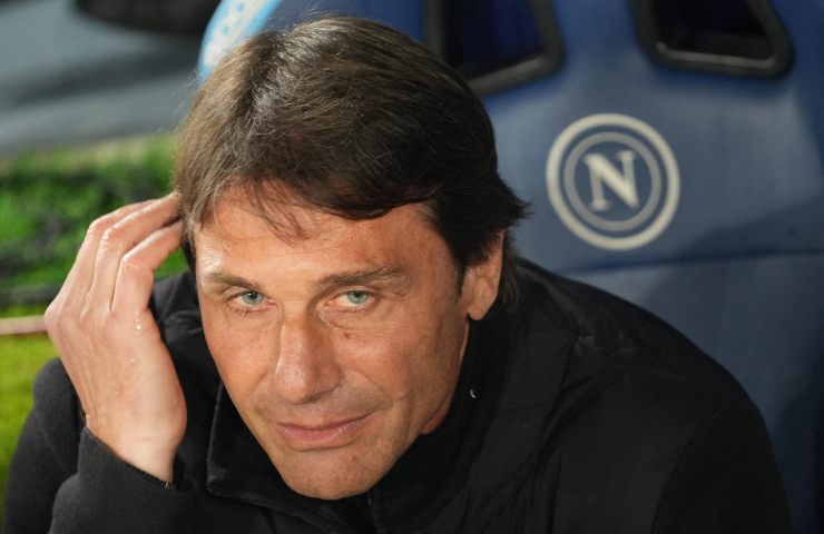 Antonio Conte in primo piano seduto in panchina con il logo del Napoli su poltroncina azzurra sullo sfondo.