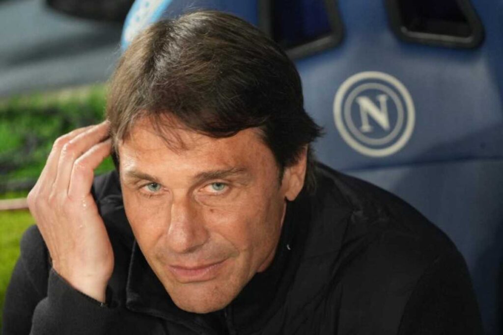 Antonio Conte sorride in panchina con il logo del Napoli in secondo piano.