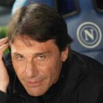 Antonio Conte sorride in panchina con il logo del Napoli in secondo piano.