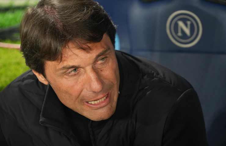 Antonio Conte seduto in panchina con il logo del Napoli sullo sfondo.