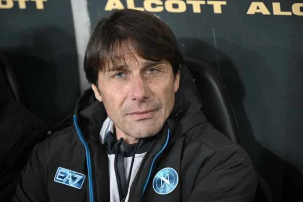 Antonio Conte con la tuta di rapprsentanza del Napoli seduto in panchina mentre guarda verso l'obiettivo.
