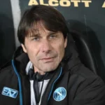 Antonio Conte con la tuta di rapprsentanza del Napoli seduto in panchina mentre guarda verso l'obiettivo.
