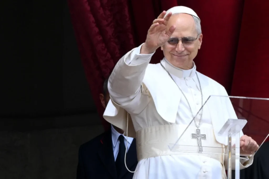 Papa Leone XIV sorride mentre saluta la folla davanti a sé col braccio destro alzato.