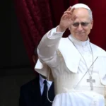 Papa Leone XIV sorride mentre saluta la folla davanti a sé col braccio destro alzato.