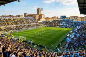 Stadio Ennio Tardini di Parma gremito per la partita di Serie A Parma-Napoli: tifosi del Parma in gialloblù a sinistra e tifosi del Napoli in azzurro a destra si circondano la struttura e il campo da gioco.